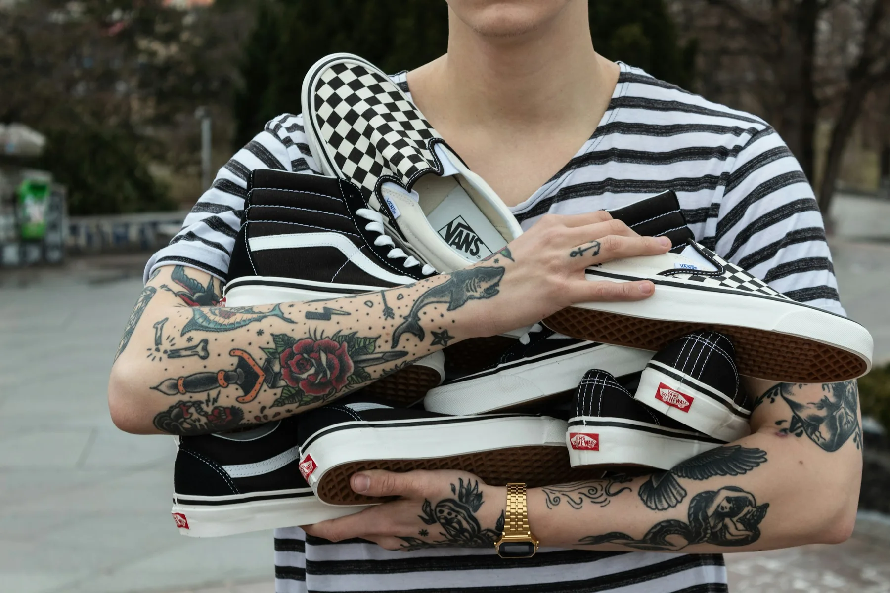 A person in a striped shirt with tattooed arms cradling an armful of black and checkerboard Vans skate shoes