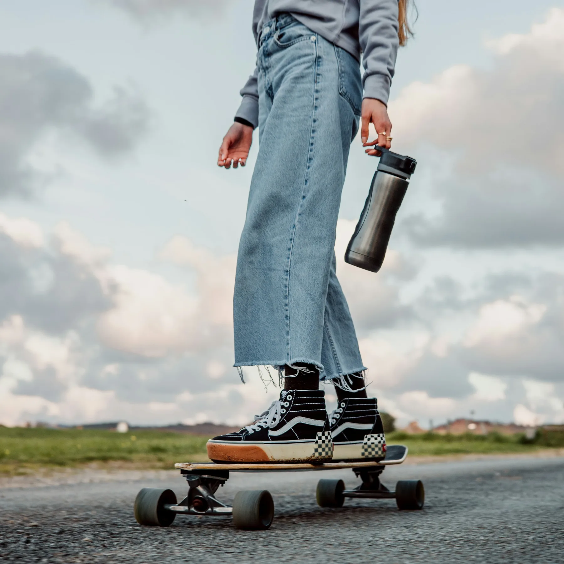 A skater in cropped jeans standing on a longboard on open road, wearing black high-top Vans