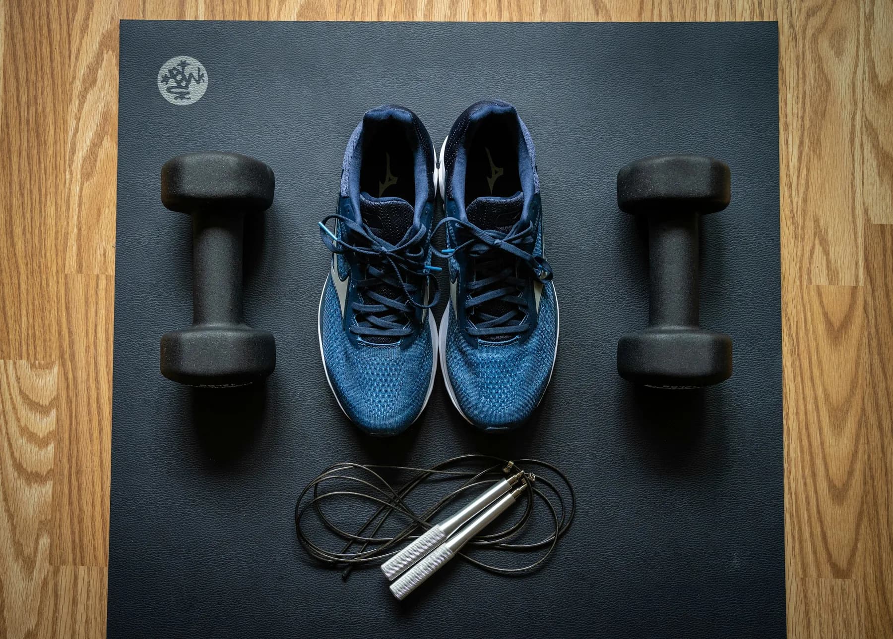 A pair of blue running shoes laid out on a dark exercise mat between two dumbbells, with a skipping rope coiled below them
