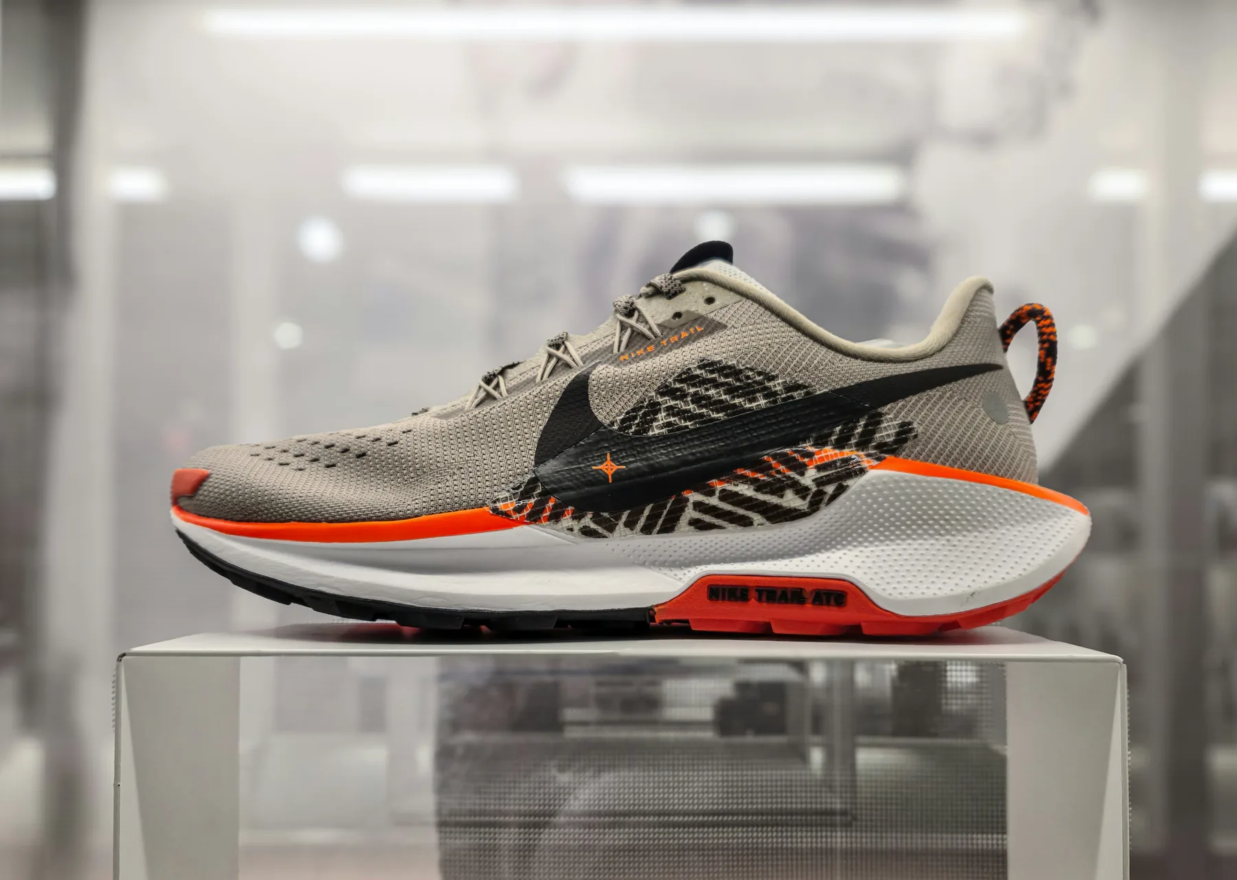 A grey trail running shoe with an orange midsole stripe, shown in profile on a lit display plinth